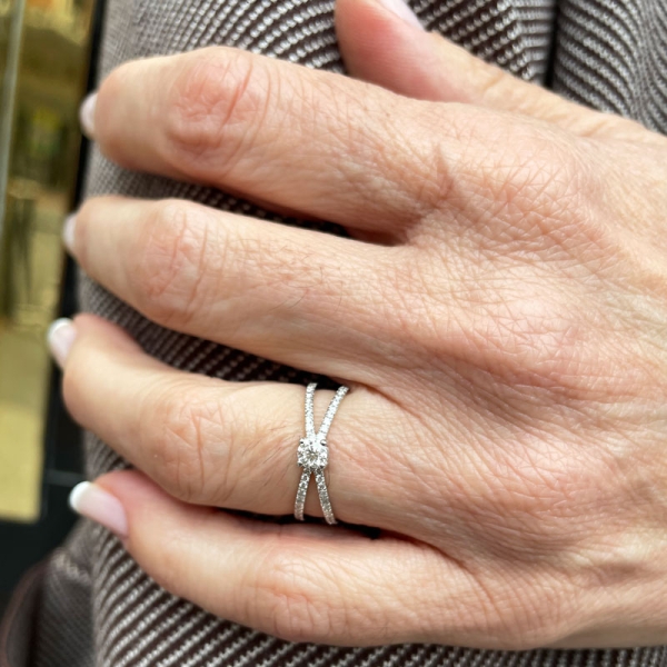 Solitario cruzado con un diamante central en talla brillante y dos brazos a los lados que se unen en el centro. En oro blanco de