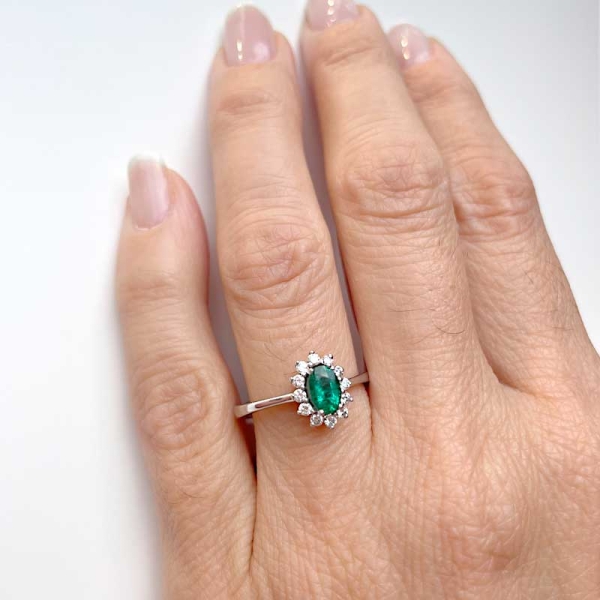 Sortija de estilo clásico con una esmeralda central en talla oval y orla de diamantes alrededor. Oro blanco de primera ley